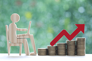 wooden man sitting by desk stack of coins with red arrow pointing up