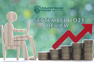 Assured Soccer Profits September 2023 results wooden man by desk red arrow on pile of coins
