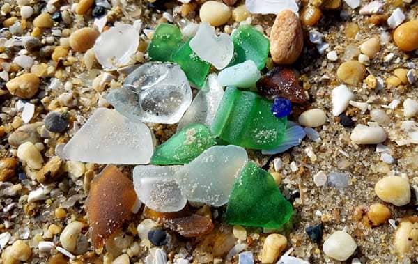 Many pieces of green, blue, brown and white sea glass laying on the sand at the beach.