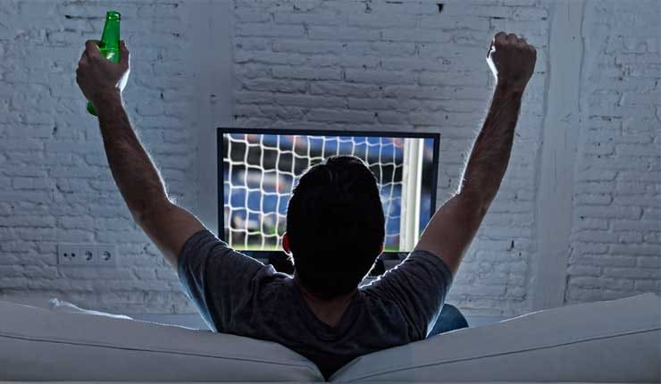 A man holding a bottle of beer, watching a football match on television and celebrating a goal