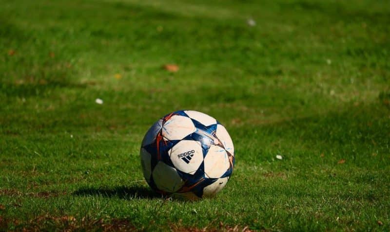 An Adidas branded football sits on a grass football pitch.